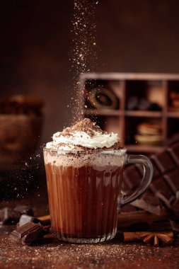 Üzerine krem şanti ve çikolata parçacıkları serpiştirilmiş sıcak çikolata. Eskimiş kahverengi arka planda yer alan malzemeler içeren bir çikolata içeceği..