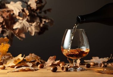 Brendi şişeden bardağa dökülür. Kurumuş meşe yapraklarıyla ahşap bir masada konyak..