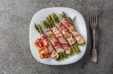 Domuz pastırmasına sarılı ızgara yeşil kuşkonmaz. Fotokopi alanı olan gri taşlı arka planda beyaz tabakta ev yapımı aperatif. Üst görünüm.