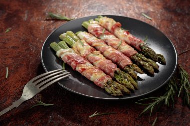 Izgara yeşil kuşkonmaz. Siyah tabakta domuz pastırması. Kahverengi bir arka planda..