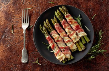 Domuz pastırmasına sarılı ızgara yeşil kuşkonmaz. Kahverengi arka planda siyah tabakta ev yapımı aperatif. Üst görünüm.