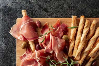 Tahta kesme tahtasında ekmek çubuğu, zeytin ve biberiyeli jambon. Geleneksel İtalyan atıştırmalıkları siyah taş bir masada..