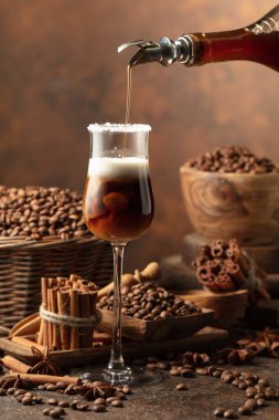 Kahve likörü bir şişeden kremalı bir bardağa dökülür. Kahve çekirdekleri, tarçın çubukları ve anason masaya saçılmış..