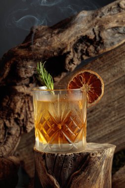 Eski usul bir kokteyl, buzlu, tahta bir kütükte. Kurutulmuş portakal dilimi ve yanan biberiye filizi ile süslenmiş soğuk, ferahlatıcı bir içecek..