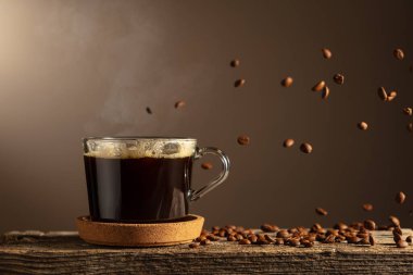 Eski bir tahtanın üzerinde cam bardakta sade kahve. Kavrulmuş kahve çekirdekleri hareket halinde. Boşluğu kopyala.