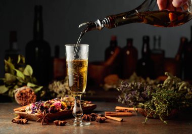 Sarı bitkisel likör ya da karışım eski bir şişeden bardağa dökülür. Kurutulmuş otlar, çiçekler, baharatlar ve eski mutfak aletleri. Bitkisel ilaç kavramı.