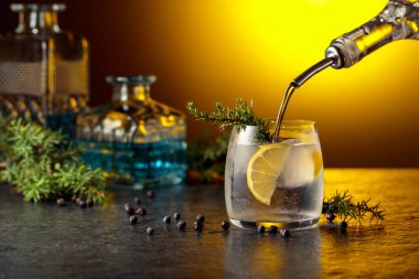 Kokteyl cini ve tonik, ardıç dalları ve siyah bir masada limon dilimleri. Cin şişeden buz ve limon dilimli bardağa dökülür. Sarı arka ışık.