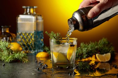 Kokteyl cini ve tonik, ardıç dalları ve siyah taş bir masada limon dilimleri. Kokteyl bir karıştırıcıdan buzlu bir bardağa dökülür. Sarı arka ışık.