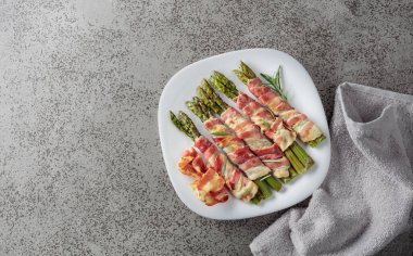 Domuz pastırmasına sarılı ızgara yeşil kuşkonmaz. Fotokopi alanı olan gri taşlı arka planda beyaz tabakta ev yapımı aperatif. Üst görünüm.