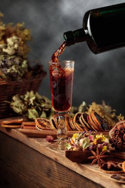 Bitkisel merhem ya da tentür eski bir şişeden bardağa dökülür. Bitki gibi doğal malzemeler, kurutulmuş turunçgil dilimleri, görünür baharatlar. Aromatik sıvı ev yapımı, sanatsal bir zanaat öneriyor.