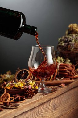 Bitkisel merhem ya da tentür eski bir şişeden bardağa dökülür. Bitki gibi doğal malzemeler, kurutulmuş turunçgil dilimleri, görünür baharatlar. Aromatik sıvı ev yapımı, sanatsal bir zanaat öneriyor.