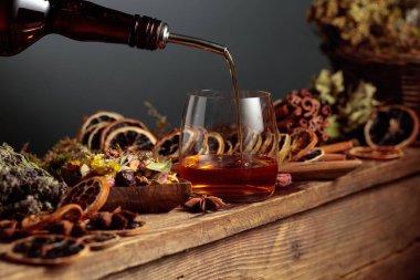 Bitkisel likör eski bir şişeden bardağa dökülür. Bitkiler, kökler, kurutulmuş narenciye dilimleri, görünen baharatlar gibi doğal malzemeler. Aromatik kehribar sıvısı ev yapımı bir zanaat olduğunu gösteriyor..