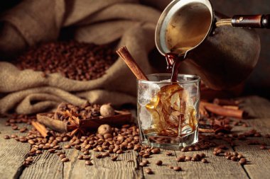 Buzlu kahve hazırlığı. Türk bakır kahve demliğinden buzlu bir bardağa sade kahve dökülüyor. Eski ahşap bir masa üzerinde kavrulmuş kahve çekirdekleri bir çanta yatar ve masaya yayılmış tarçın çubukları, yıldız anason ve kurutulmuş narenciye dilimleri vardır.