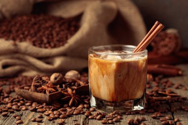 Kremalı buzlu kahve. Eski ahşap bir masa üzerinde kavrulmuş kahve çekirdekleri bir çanta yatar ve masaya yayılmış tarçın çubukları, hindistan cevizi ve anason yıldızları vardır..