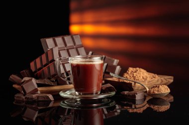 Bir kupa sıcak çikolataya karşı kırık çikolata ve kakao tozlu ahşap tabak. Siyah parlak arka plan derinlik katıyor. Boşluğu kopyala.