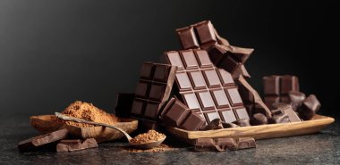 Tahta bir kase kakao tozunun yanında koyu çikolata kırıntıları. Siyah arka plan. Kakao tozlu bir kaşığa odaklan. Boşluğu kopyala.