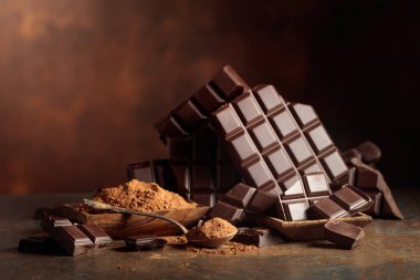 Tahta bir kase kakao tozunun yanında koyu çikolata kırıntıları. Sıcak kahverengi arka plan çikolata renginin zenginliğini vurgular. Boşluğu kopyala.