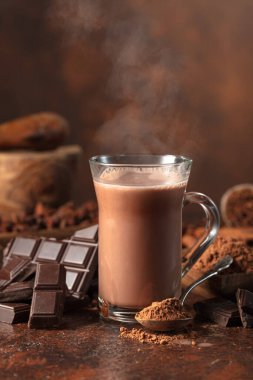 Bir parça acı çikolata, kakao tozlu kaşık ve kahverengi bir masada bir bardak kakao..