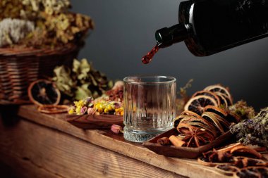 Bitkisel merhem ya da tentür eski bir şişeden bardağa dökülür. Bitki gibi doğal malzemeler, kurutulmuş turunçgil dilimleri, görünür baharatlar. Aromatik sıvı ev yapımı, sanatsal bir zanaat öneriyor.