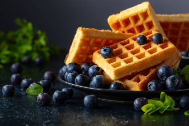 Siyah tabakta taze yaban mersinli Belçika waffleları. Koyu mavi bir masada yeşil yapraklı waffle ve taze meyveler..