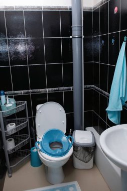 Tuvaletli bir dolap, çocuklar için mavi bir koltuk, bir havlu, gri bir kanalizasyon borusu, plastik bir raf, siyah fayanslar ve bir çöp kutusu..