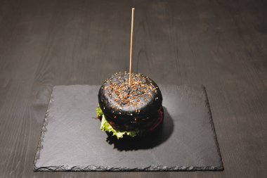 Düz siyah tabakta ev yapımı burger. Susamlı esmer çörek, peynirli köfte, marul, soğan, domates..