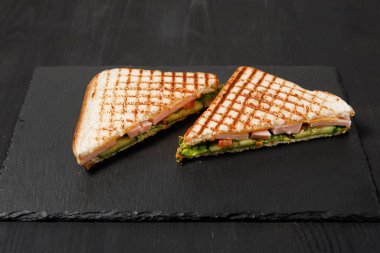 Düz tabakta üç köşeli ev yapımı sandviç. Tost, salata, tavuk, domatesler siyah bir masada..