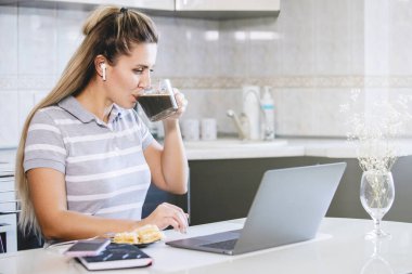 Kadın kahve içiyor ve masada dizüstü bilgisayarla çalışıyor. Tabakta pasta, günlük, bardak ve akıllı telefon var. Seçilmiş Odak. Coronavirus karantina ödevi, sosyal mesafe, özel girişimci.