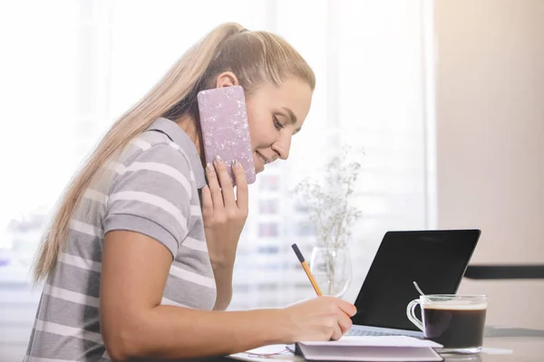 Woman listens to a smartphone and takes notes at homework. Cup of coffee and laptop on the table. Social distance. Epidemic, coronavirus pandemic. Side view.