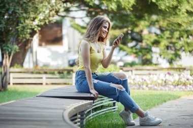 Kadın, şehir parkındaki bankta sarı tişörtlü ve kot pantolonlu bir akıllı telefonla mesajlar okuyor..