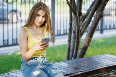 Kadın, şehir parkındaki bankta sarı tişörtlü ve kot pantolonlu bir akıllı telefonla mesajlar okuyor..