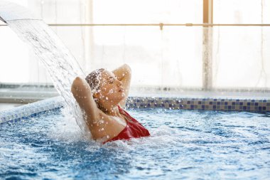 Havuzdaki su deresinin altında kırmızı mayolu sarışın bir kadın. Otel spa çeşmesine mavi su dökülüyor..