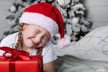 Kırmızı şapkalı, Noel hediyeli bir kız. Yatağa uzanır, kutuya sarılır ve Noel ağacının arka planına yaslanır..