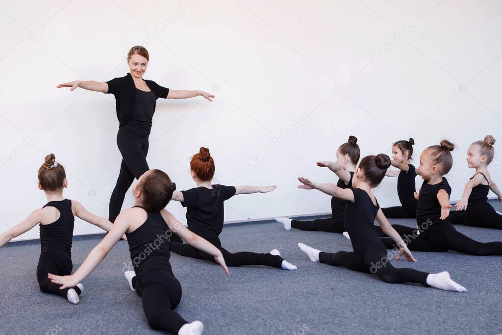 Girls do gymnastic split under the guidance of a coach in dance training. Black leotard, hair in a bun, white socks.