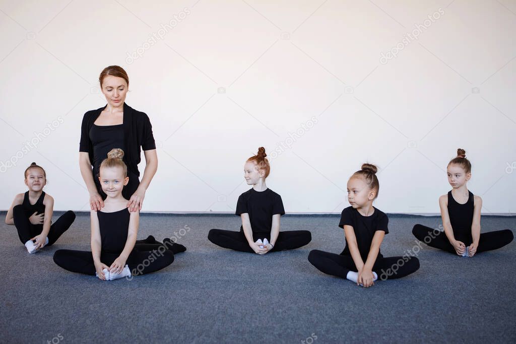 Girls gymnasts are sitting in a lotus position at a warm-up under the guidance of a trainer in dance training. Black leotard, hair in a bun, white socks.