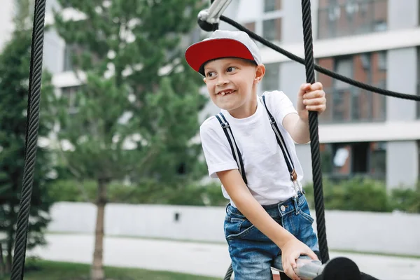 少年は市内の遊び場でロープ機器に登る レンガ造りの家の庭にサスペンダー付きの白いシャツ キャップ デニムのショートパンツ — ストック写真