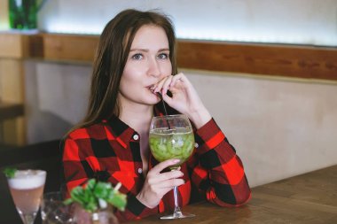 Kareli gömlekli bir kadın restorandaki bir masada bardaktan kokteyl içiyor..