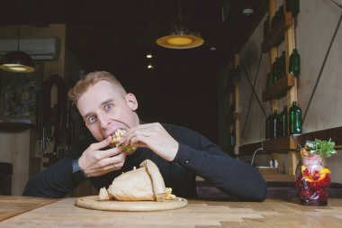 Genç sarışın adam kafedeki bir masada sandviç yemekten hoşlanıyor..