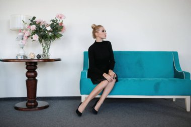 Güzel sarışın kanepede oturuyor, saçları topuz şeklinde, siyah elbise içinde, zarif bir tarzda..