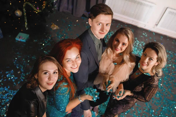 Group of friends celebrates the New Year with confetti near the Christmas tree. People laugh and rejoice.