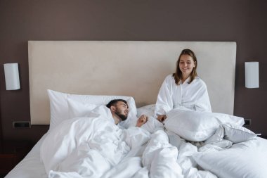 Beyaz yataklı bir kadın ve uyuyan sakallı bir adam. Otelde dinlenir, turistleri gezeriz..