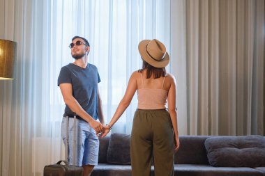 Şapkalı ve güneş gözlüklü bir adam ve kadın perdeli bir penceresi ve kanepesi olan bir otel odasına girdiler..