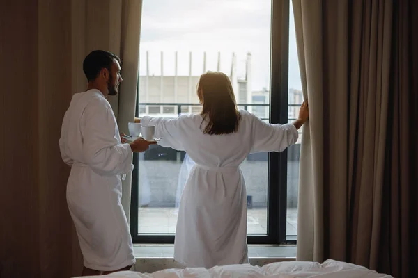 一个男人和一个女人穿着白色长袍 在卧室的窗户边喝咖啡 — 图库照片