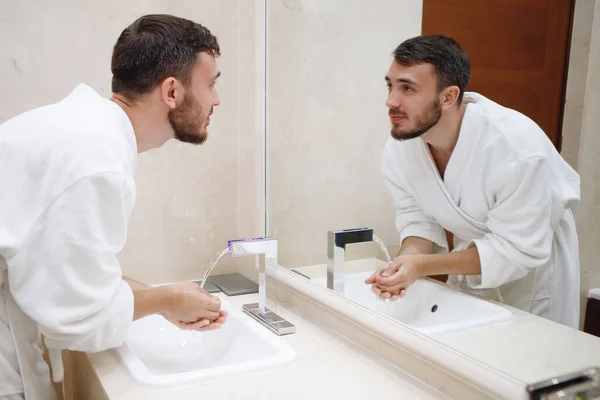Bearded guy in a white robe washes himself in the sink with a tap in the bathroom.
