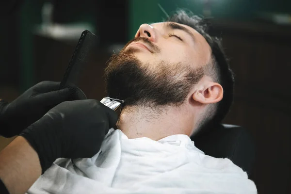 Master cuts a guy\'s beard with a hair clipper in a hairdressing chair in a barbershop salon. Selective focus, blurred background.