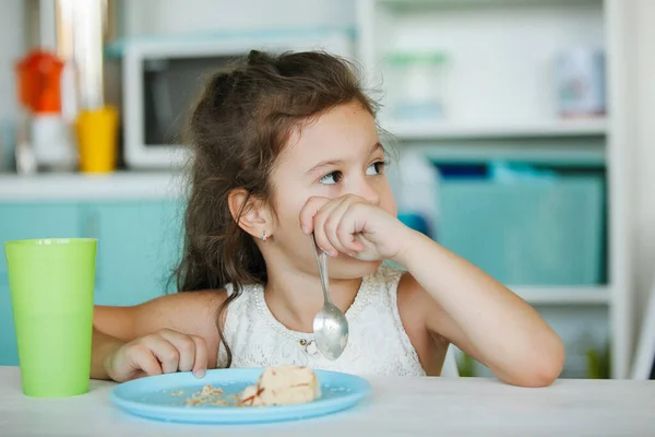 Cute girl is eating a cake. On the table is a blue plate, a green glass, holds a spoon in his hand. Family happiness, happy smile, children\'s holiday.
