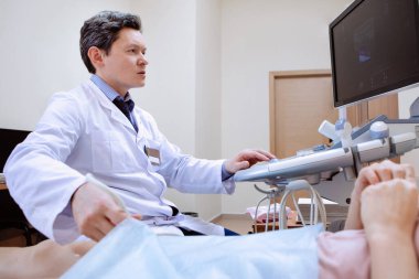 Bacakların ultrason muayenesi. Doktor genç bir kadının kan damarlarını tarıyor. Koltuk, monitör ve sinyal sensörü olan modern cihazlar, tıbbi klinik..