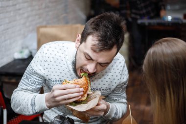 Genç adam kafede bir masada hamburger yiyor.