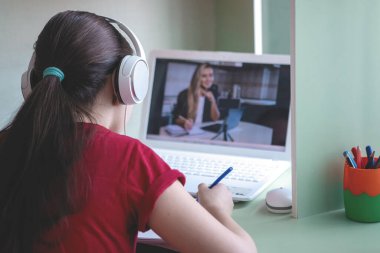 Kulaklıklı genç kız Coronavirus salgını sırasında evde dizüstü bilgisayarla ders çalışıyor. Bir deftere yazıyor. Evde eğitim. Sosyal uzaklık. Çevrimiçi okul.