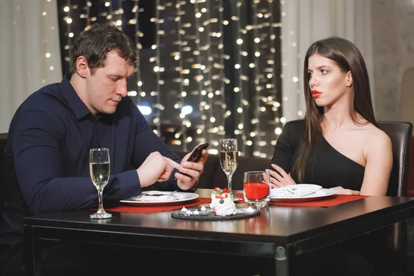Conflict of a couple at a table in a restaurant, a man looks at a smartphone, a woman is annoyed.